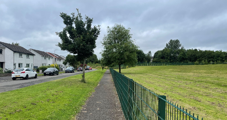Houses are on the left and a green field is on the right with a footpath in the middle.