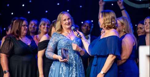 Recipient in a blue dress smiling and holding an award at a ceremony, surrounded by applauding attendees on a stage with a starry backdrop. Recipient in a blue dress smiling and holding an award at a ceremony, surrounded by applauding attendees on a stage with a starry backdrop.