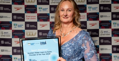 Person holding a "Sport Aberdeen Scottish Water Learn to Swim Provider of the Year 2024" award in front of a banner with various sponsors including Scottish Water and Speedo. Person holding a "Sport Aberdeen Scottish Water Learn to Swim Provider of the Year 2024" award in front of a banner with various sponsors including Scottish Water and Speedo.