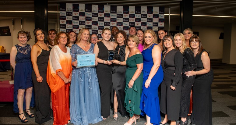 Group of individuals holding an award at a formal event, posing in front of a banner with logos. Group of individuals holding an award at a formal event, posing in front of a banner with logos.