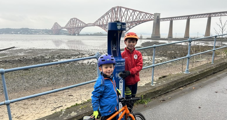 Thomas and Murray beside South Queensferry Top Up Tap Two young children pose beside bright blue water refill fountain with several large bridges in the background