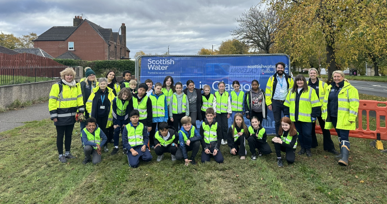 Students with the project team planting bulbs Group of people of various ages wearing safety vests, standing in front of a Scottish Water truck, in a suburban area.