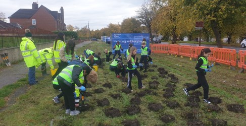 Glasgow Resilience Project team planting bulbs in Mosspark Group of volunteers wearing safety vests planting trees by the roadside, with a sign reading "Investing in Glasgow".
